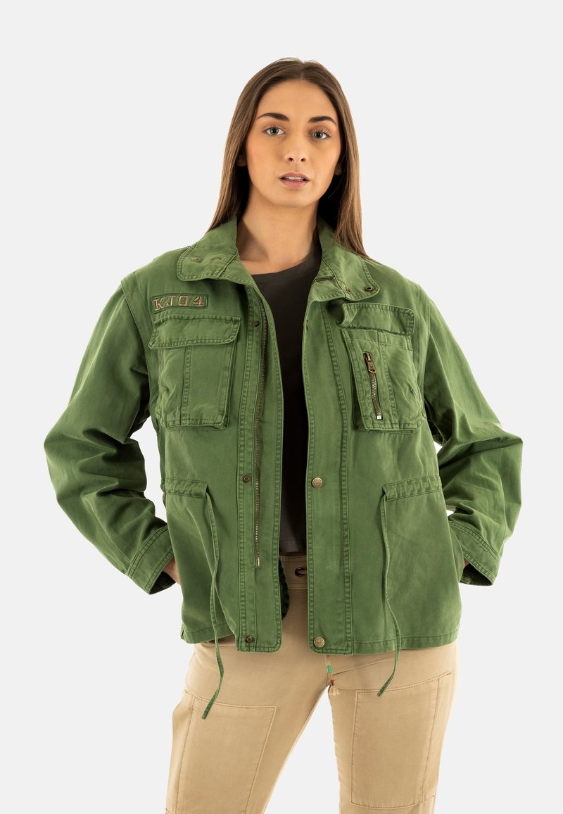 Chaqueta de algodón verde con múltiples bolsillos frontales, cierres con cremallera y a presión, dobladillo ajustable con cordón y un sutil bordado en el cuello.