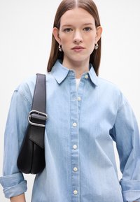 Világoskék farmering klasszikus gallérral és fehér gombokkal, fekete bőr táskával párosítva, amely ezüst csatós pánttal rendelkezik.