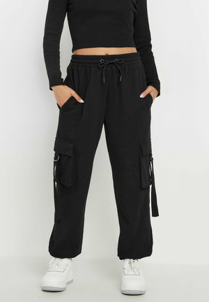 Osoba nosząca czarne spodnie typu cargo z troczkiem w talii, ręce w kieszeniach, połączone z białymi sneakersami na platformie i czarnym krótkim swetrem.