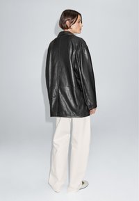 Černá kožená oversized bunda s klopovým límcem a viditelnými švy, doplněná volnými krémovými kalhotami. Pohled zezadu.