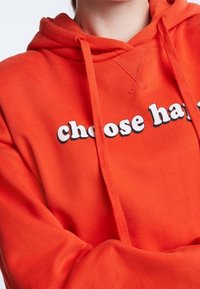 Sudadera naranja con cordones, que presenta el texto "elige ser feliz" en letras blancas y en negrita a lo largo del pecho. Textura de tela suave.
