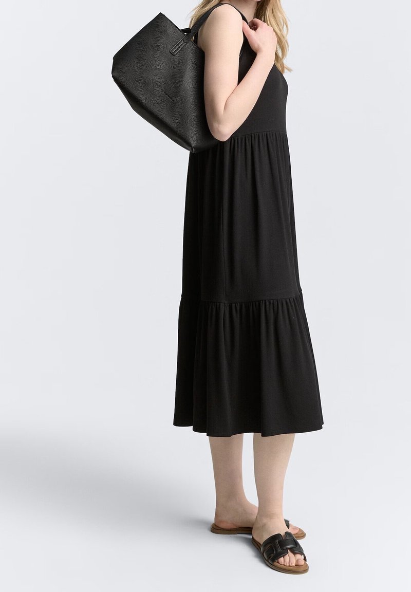 Femme portant une robe midi noire sans manches et des sandales noires à enfiler, portant un grand sac à épaule noir texturé.