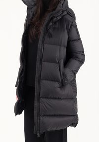 Zwarte pufferjack met capuchon, voorzien van doorgestikte panelen, een rits aan de voorkant en zijzakken. Het materiaal is glanzend en soepel.