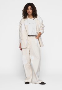 Blazer oversize color beige claro con grandes patrones florales blancos, combinado con pantalones de pierna ancha a juego y una camiseta blanca lisa, acentuado con un cinturón marrón.