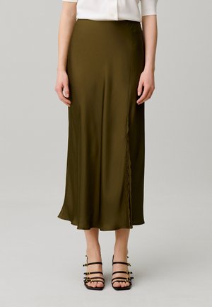 Femme portant une jupe midi vert olive avec une fente latérale et des sandales à talons hauts noires à brides, debout devant un fond uni.
