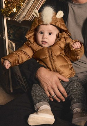 Braune Steppjacke mit Kapuze, die weiße Kunstpelzbesätze und Ohrenakzente aufweist. Das Baby trägt graue gemusterte Hosen und beigen Schuhe. Die Hände sind sichtbar.