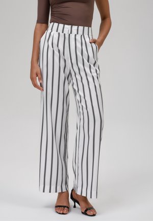 Žena nosící bílé široké kalhoty s vertikálními černými pruhy, hnědý přiléhavý top a černé boty s otevřenou špičkou, stojící s rukama v kapsách.