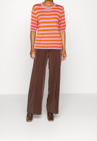Maglione a righe in arancione e lilla, maniche corte, scollatura rotonda, abbinato a pantaloni larghi marroni e stivaletti marroni.