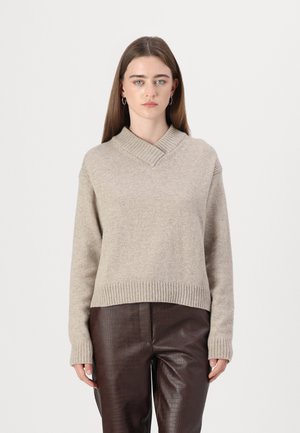 Beżowy sweter dzianinowy z dekoltem w serek i prążkowanym wykończeniem, w zestawieniu z brązowymi skórzanymi spodniami z teksturą krokodyla.