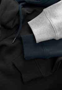 Tres sudaderas con capucha: negra, verde azulado oscuro y gris claro. Cuentan con puños acanalados, cordones y una textura suave y lisa.