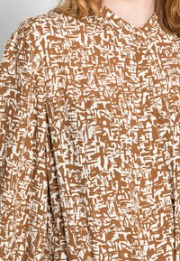 Braun-weißes, abstraktes gemustertes Langarmhemd mit einem kleinen Knopf am Kragen, getragen von einer Person mit heller Haut und rötlichem Haar.