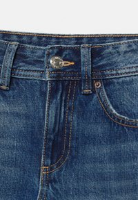 Denim kavbojke v temno modri barvi, klasičnega pet-džepnega designa, vidno šivanje in srebrn gumb na pasu.