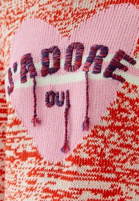 Gestrickter roter und weißer Pullover mit einem großen pinken Herz und mehrfarbigem Garntext mit der Aufschrift "HOLD ON" sowie hängenden Fransen.