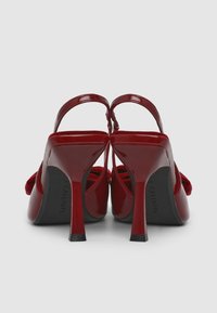 Rote Lackleder-High Heels mit offener Ferse, ausgestattet mit einem geschwungenen Riemen und einem 10 cm hohen Stiletto-Absatz. Strukturierte schwarze Sohle mit Markenaufdruck.