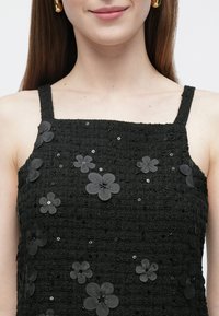 Juodas tekstūruotas viršus su kvadratiniu iškirpimu ir plonais dirželiais, papuoštas juodais gėlių aplikacijomis ir blizgučiais visame paviršiuje.