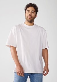 Lys grå bomulls T-skjorte med korte ermer, rund hals, løs passform og synlige sømdetaljer. Brukt sammen med blå jeans.