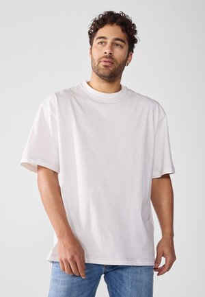Svijetlosivi pamučni T-shirt s kratkim rukavima, okruglim izrezom, ležernim krojem i vidljivim šavovima. Uparen s plavim trapericama.