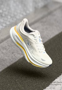 Sapatilha de corrida beige claro com sola grossa branca e amarela, com a marca HOKA, atacadores atados, mostrada no ar acima de uma superfície de estrada texturizada.