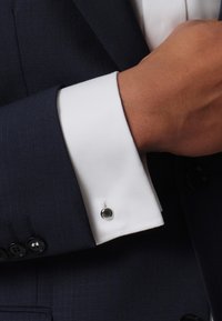 Navy suit jacket with a white cuff showing a buttonhole and a round cufflink. Textured fabric with a subtle grid pattern on the jacket.