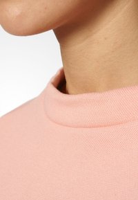 Primer plano de un cuello y línea de mandíbula que lleva un suéter de punto rosa de alto cuello contra un fondo blanco.