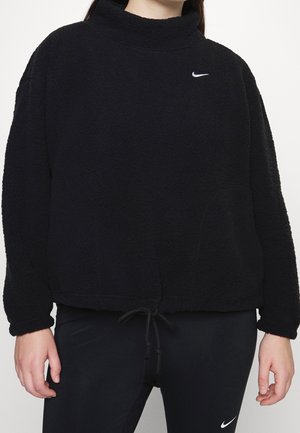 Osoba nosząca czarny polarowy sweter z wysokim kołnierzem i suwakiem na dole, w połączeniu z czarnymi legginsami Nike z białymi logo Swoosh.