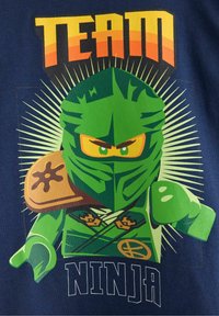 Grafica di un personaggio ninja verde su una maglietta blu navy. Presenta occhi gialli, una spallina beige e testo in grassetto che recita "TEAM NINJA."