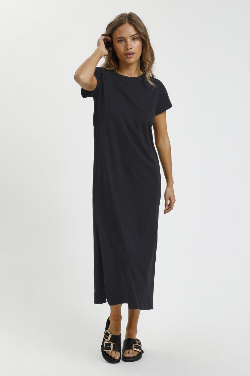 Femme debout en robe midi noire à manches courtes et sandales noires à boucle devant un fond blanc uni.