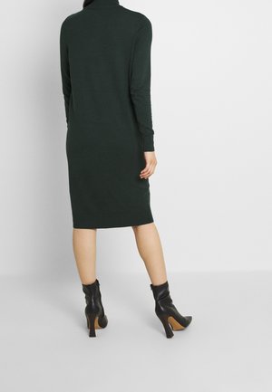 Robe tricotée vert foncé avec un col montant, des manches longues et une silhouette ajustée, se terminant juste en dessous du genou, associée à des bottines noires.
