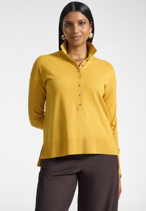 Donna con capelli neri lisci che indossa orecchini d'oro, una collana a catena dorata, un maglione giallo senape con bottoni e pantaloni marrone scuro.