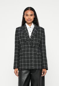Dvostruki blejzer u crno-bijelom karo uzorku, s oštrim reverima, dva prednja džepa i gumbima. Uparen s bijelom košuljom.