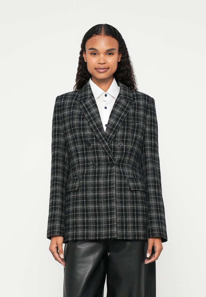 Dvostruki blejzer u crno-bijelom karo uzorku, s oštrim reverima, dva prednja džepa i gumbima. Uparen s bijelom košuljom.