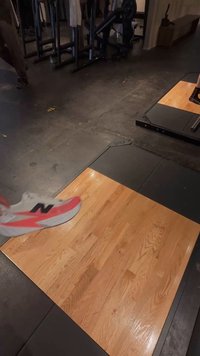 Saltea de gimnastică din lemn cu suprafață netedă, înconjurată de margini din cauciuc negru. Un picior încălțat cu un sneaker gri cu un accent roz apare deasupra.