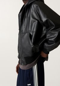 Persona che indossa una giacca in pelle nera con zip sopra una maglietta grigia e pantaloni da ginnastica blu navy con strisce bianche e logo sul lato.