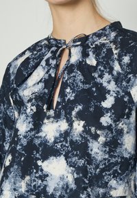 Blusa azul-marinho com um padrão branco semelhante a nuvens, apresentando um detalhe de laço no decote e um corte confortável com acentos pregueados.