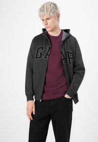 Jeune personne avec des cheveux courts blonds, portant un sweat à capuche GAP gris foncé sur une chemise bordeaux et un pantalon noir, se tenant avec une main dans la poche.