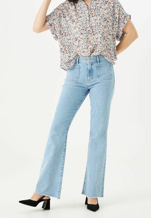 Himmelblaue, hoch taillierte Jeans mit ausgestellten Beinen, kombiniert mit einer kurzärmligen, mehrfarbigen Bluse mit Blumenmuster und schwarzen, spitzen Pumps.