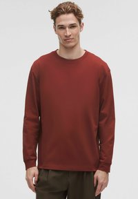 Tricou cu mânecă lungă, gât rotund, de culoare roșie închisă, realizat dintr-un material moale, cu o croială relaxată și manșete ribbate. Detalii de design simple, minime.