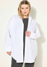Hvid oversized skjorte med knaplukning fortil, lange ærmer og buet kant. Bæres over en mørk, ribbet top med en glat stofstruktur.