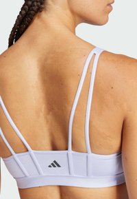 Sujetador deportivo de lavanda claro con tirantes finos ajustables, tejido texturizado y un pequeño logo negro en la parte posterior.