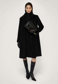 Černý vlněný kabát s přizpůsobeným designem, v kombinaci s odpovídajícím rolákem a koženými rukavicemi. Modelka má na sobě černé kozačky na vysokém podpatku, sahající až ke kolenům.