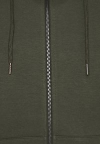 Sudadera con cremallera en color verde oliva, hecha de una tela suave con una textura lisa. Cuenta con una cremallera plateada y cordones ajustables en la capucha.