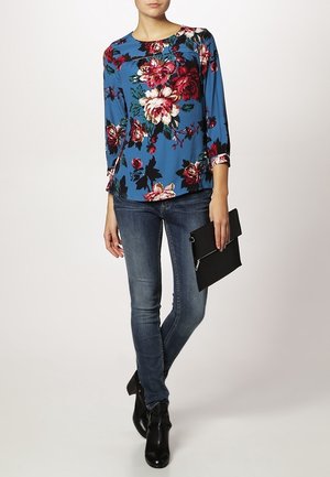 Blusa blu a stampa floreale con maniche a tre quarti, abbinata a jeans skinny blu scuro e stivaletti neri, mentre tiene una clutch nera.