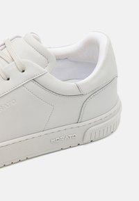 Zapatilla blanca hecha de material sintético liso con costuras tonales, cordones planos, collar acolchado y suela de goma texturizada con marca.