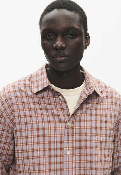 Personne noire aux cheveux courts portant une chemise blanche sous une chemise à carreaux orange et blanche à boutons, regardant droit devant avec une expression neutre.
