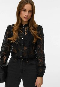 Sort bluse i blonder med lange ærmer, høj hals og perleknapdetaljer, kombineret med højttaljede sorte jeans og en sort håndtaske.