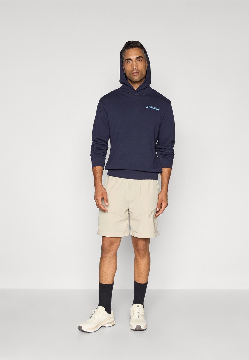 Felpa con cappuccio blu marina con logo blu; abbinata a pantaloni corti beige, calze scure e scarpe da ginnastica bianche con dettagli grigi.