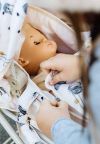 Bambola in un passeggino di stoffa rosa con un copertura a motivo floreale. La bambola ha i capelli biondi e una carnagione neutra, gli occhi chiusi e una bocca leggermente aperta.