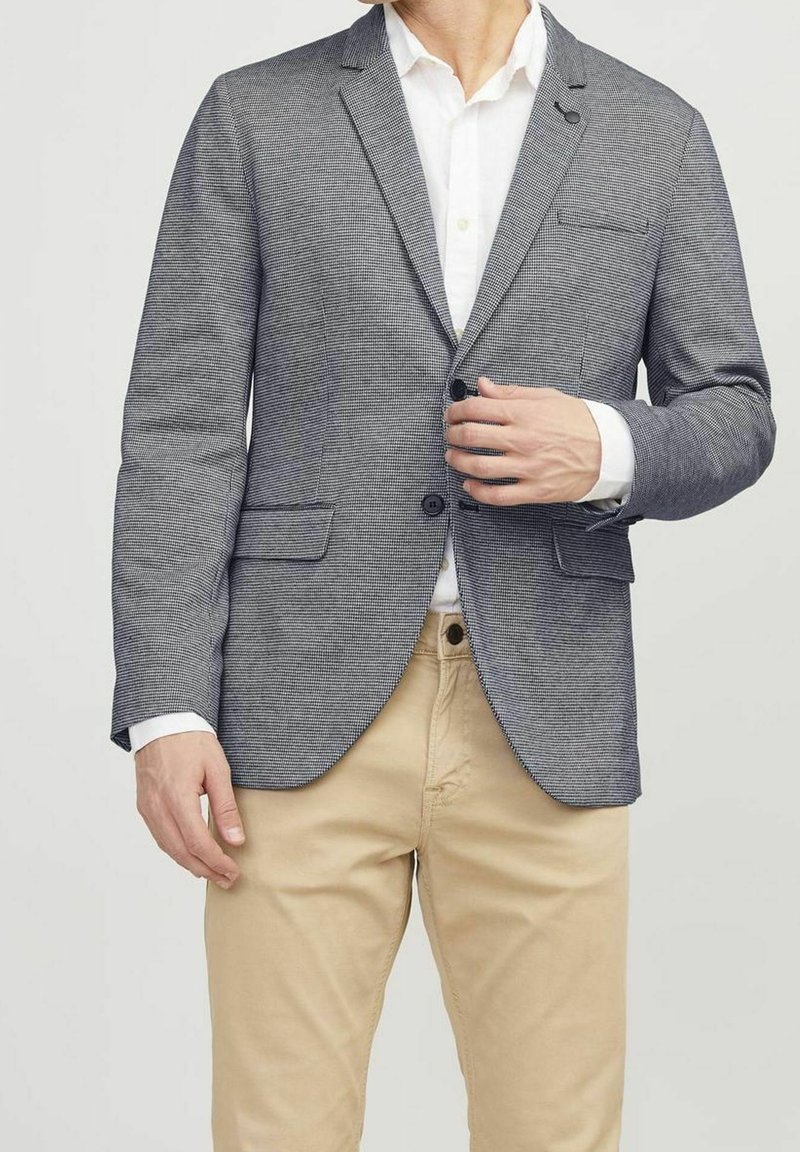 Grau karierter Blazer mit Aufschlagkragen, einem Knopfverschluss und zwei Vorderwassertaschen. Getragen über einem weißen Hemd und beigen Hosen.