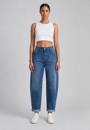 Vrouw staat en draagt een wit cropped tanktop, losse blauwe high-waisted jeans met omgeslagen pijpen, en witte sneakers op een grijze achtergrond.