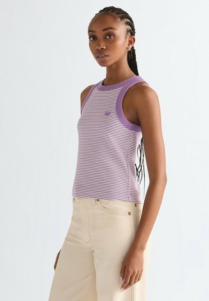 Giovane donna con capelli intrecciati che indossa un top senza maniche a righe viola e bianche e pantaloni beige chiaro, in piedi di lato su uno sfondo semplice.